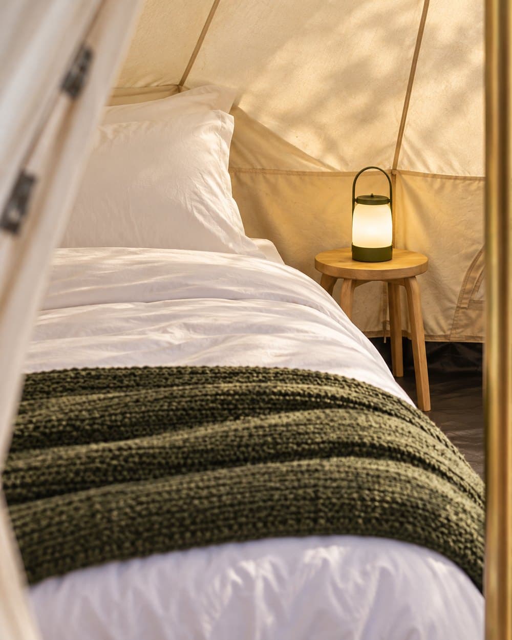 Illustrative close-up of white bed linen, an olive throw and a warm lantern against cream canvas.